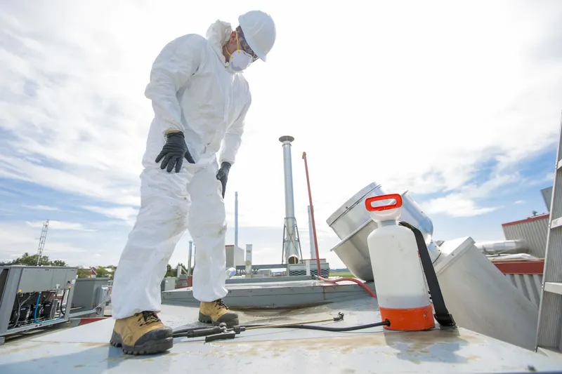 Two technicians on a flat commercial roof at dusk beside an upblast exhaust fan, with low-rise plaza buildings and a highway in the distance