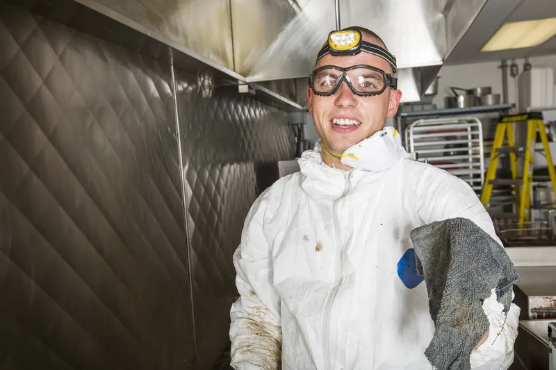 Technician in protective gear degreasing the wall and floor behind a pulled-out fryer bank on a commercial kitchen cook line