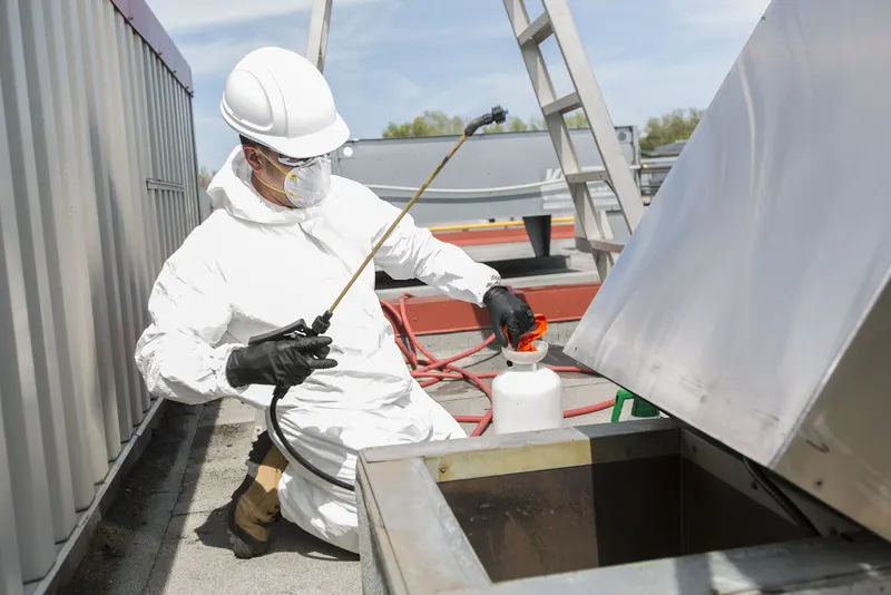 Technician kneeling beside a rooftop upblast exhaust fan tipped open on its hinge kit, with the blower wheel and curb exposed