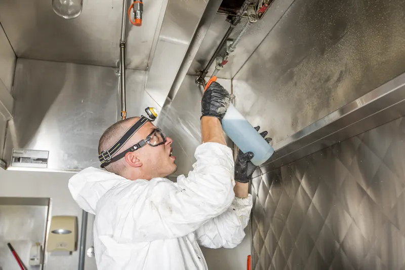 Technician spraying degreaser into the underside of a stainless steel canopy hood above a restaurant cook line, with filters removed