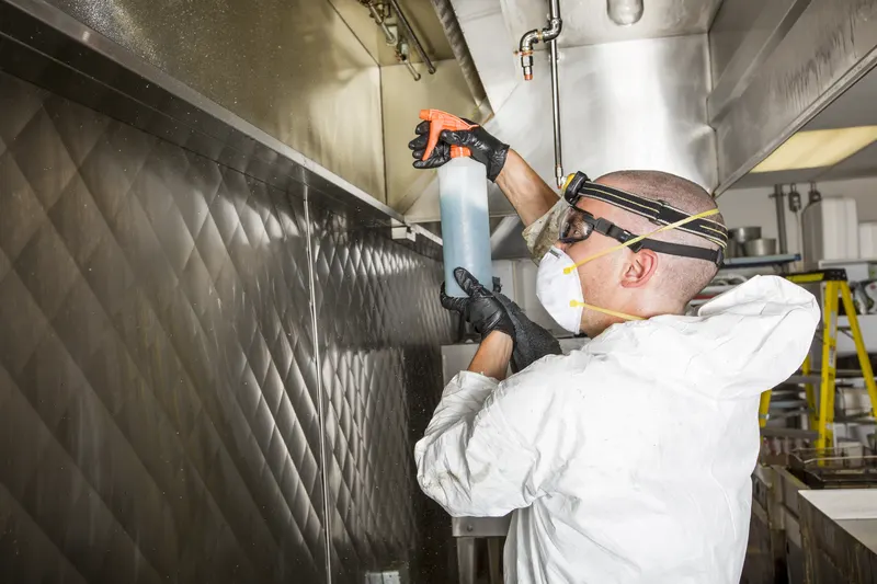 Technician spraying degreaser across the underside of a stainless steel commercial kitchen canopy with the baffle filters removed