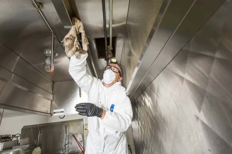 Gloved hand wiping the inside wall of a stainless steel hood plenum with a cloth, showing the grease film behind the filter slot